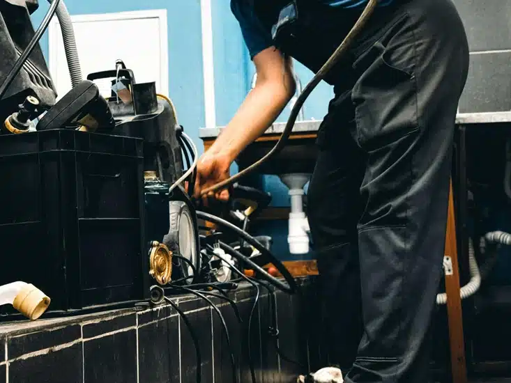 A technician in a blue shirt and black apron inspects a long hose in a maintenance workshop with industrial equipment in the background.