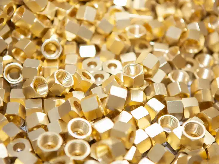 A pile of numerous shiny brass hexagonal nuts with internal threading.