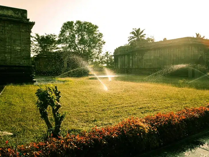 Several automated sprinklers spraying water over a wide green field between stone buildings during a golden sunset.