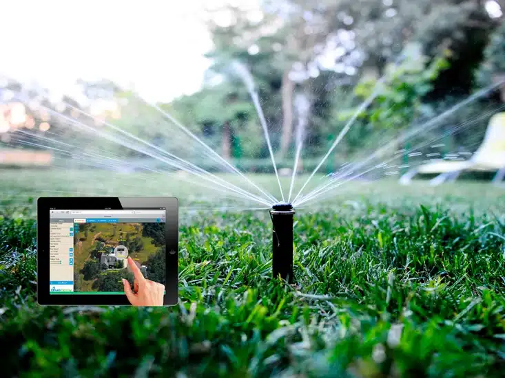 A tablet showing an irrigation control app in front of a working lawn sprinkler on a green grass field.